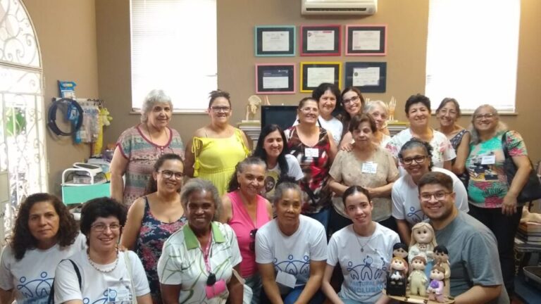 Descrição da Imagem: Fotografia de um grupo de 20 pessoas, sendo 19 mulheres e 1 homem. Eles possuem diferentes idades, alguns estão abaixados na frente dos outros e sorriem. O homem está agachado segurando uma imagem feita em feltro de Nossa Senhora do Amor Inclusivo. Eles estão em uma sala com paredes marrom, na parede do fundo há um ar condicionado, seis quadros coloridos com diplomas entre duas janelas. Na lateral esquerda há uma estante verde ao lado de uma porta de vidro. Fim da descrição.